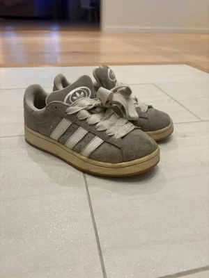 Grå Adidas Campus sneakers i mocka - Säljer ett par grå Adidas Campus sneakers i mocka med klassiska vita ränder på sidorna och vit logga på plösen och hälen. Skorna har breda vita snören och en beige sula. Perfekta för dig som gillar retrostil och streetwear. (JAG rengör såklart dom med en mockaborste och sluorna, innan dom sickas)!!!!!!!!!!!