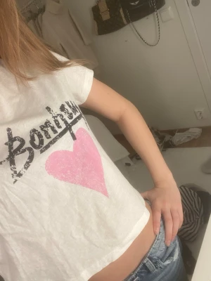 Vit croppad t-shirt med tryck - Säljer en vit croppad t-shirt med svart texttryck och ett stort rosa hjärta på framsidan. T-shirten har korta ärmar och är i en loose passform, perfekt till jeans eller shorts. Materialet känns som bomull och är mjukt mot huden.