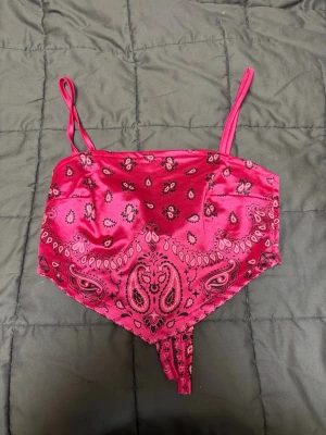 Rosa bandana-väst från FB Sister - Säljer en rosa väst från FB Sister i bandana-stil med tunna axelband och knytning i ryggen. Västen har ett paisleymönster och är tillverkad i ett glansigt, mjukt material, storlek XXS