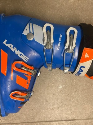 Blå Lange RSJ 60 pjäxor strl 26.5 och 308 mm - Säljer ett par blå Lange RSJ 60 pjäxor med orangea detaljer och fyra metallspännen. Insidan är svart för extra stilpoäng. Perfekta för dig som vill ha stabilitet och cool design i backen. Skalet är i slitstark plast och innerskon är mjuk och bekväm.
