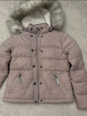 Ljusrosa pufferjacka från Belle Amie - Säljer en ljusrosa pufferjacka från Belle Amie med mysig fuskpäls på huvan och vitt teddyfoder inuti. Jackan har dragkedja, knappar framtill och två snedställda fickor med dragkedja. Perfekt för kalla dagar och riktigt skön att bära. I barnstorlek. Är i väldigt bra skick men har ett namn inuti lappen.