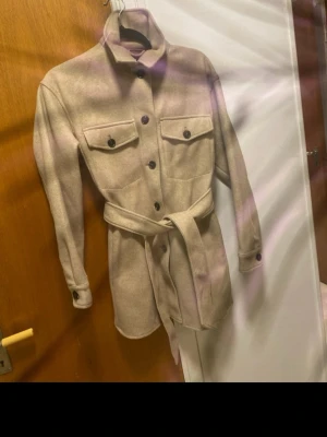 Beige overshirt från Zara med bälte - Säljer en beige overshirt från Zara i ullmix med knappar framtill, två bröstfickor och bälte i midjan. Jackan har lång ärm med knapp vid ärmslut och en clean, rak passform. Perfekt för lager på lager och snygg till både jeans och kjol. Oanvända 