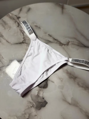 Vit bikiniunderdel Victoria's Secret - Säljer en vit bikiniunderdel från Victoria's Secret med smala axelband dekorerade med glittriga stenar. Snygg och enkel design med elastiska band som ger en bekväm passform. Perfekt för strand eller poolhäng.