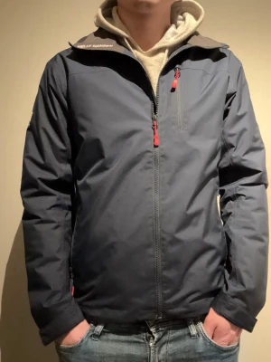 Mörkblå vindjacka från Helly Hansen - Säljer en mörkblå vindjacka från Helly Hansen med Helly Tech-tryck på ärmen. Jackan har röda dragkedjor, hög krage och flera fickor. Insidan är fodrad med mjuk fleece för extra komfort. Perfekt för blåsiga dagar och outdoor. Navy