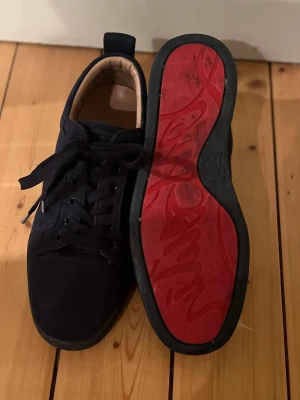 Christian Louboutin  - Snygga mörkblå sneakers i mocka med klassisk snörning och rund tå. Skorna har en unik röd sula med grafiskt mönster och svart kant. Insidan är ljusbeige och ger en schysst kontrast till utsidan. Perfekta för dig som vill ha något stilrent men ändå sticker ut.