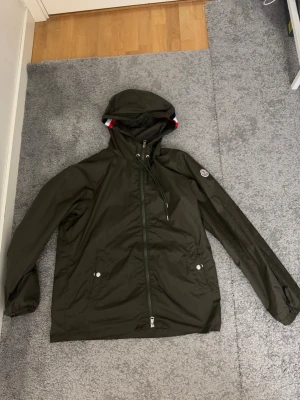 Mörkgrön Moncler vindjacka med huva - Snygg mörkgrön vindjacka från Moncler med huva och snörning. Jackan har dragkedja framtill, två fickor med tryckknappar och Moncler-logga på ärmen. Materialet är lätt och syntetiskt, perfekt för blåsiga dagar. Stilsäker och enkel att matcha med din outfit. Pris kan absolut diskuteras och storleken är Size 3 