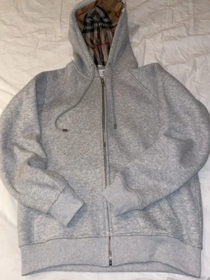  Grå Burberry Hoodie - Säljer en klassisk grå Burberry-kofta med dragkedja och huva. Använd endast en gång, så den är i väldigt fint skick. Tidlös design med Burberrys ikoniska rutmönster på insidan av huvan och diskret logotyp på bröstet. Mjuk och bekväm kvalitet, perfekt både till vardags och för en mer uppklädd casual look.    Jag är öppen för rimliga bud vid snabb affär.