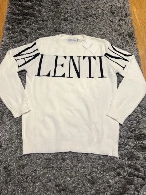 Vit tröja från Valentino - Säljer en vit stickad tröja från Valentino med svart logotyp över bröstet och coola svarta detaljer på ärmarna. Tröjan har rund halsringning och långa ärmar. Perfekt för dig som vill ha en statement piece i garderoben.