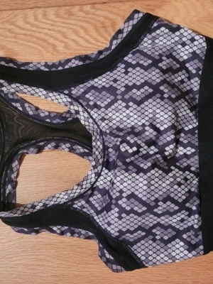 Svart och grå sport bh  med mönster - Säljer en sportig bh i svart och grått med ett coolt orminspirerat mönster. Den har meshdetaljer i ryggen för extra ventilation och breda svarta kanter. Perfekt för dig som vill sticka ut på gymmet eller under löprundan.