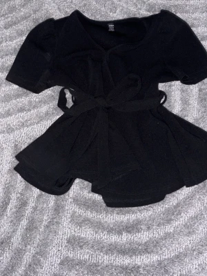 Svart omlottopp med knyt från SHEIN - Säljer en svart omlottopp från SHEIN i storlek S. Toppen har korta ärmar, markerad midja med knytband och en mjuk, följsam känsla. Perfekt för dig som gillar en enkel men snygg look.