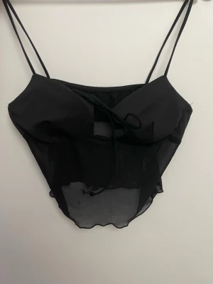 Svart meshväst med tunna band - Säljer en svart väst med tunna axelband och meshdetaljer. Västen har snörning framtill och är croppad med transparent nederdel. Perfekt för dig som vill ha en edgy och trendig look.