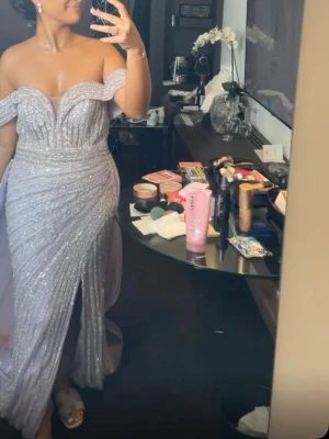 Ljuslila glittrig balklänning offshoulder - Otroligt vacker och festlig klänning i en ljus lila nyans med mycket glitter som fångar ljuset fantastiskt fint. Klänningen har en elegant off-shoulder-ringning med draperade axelband och en figurnära passform. Köpt från Catwalk Couture. Lite defekter på släpet, men inget som syns jätte mycket!   Den är vackert draperad framtill och har en hög slits. Perfekt till bröllop, bal, förlovningsfest eller andra speciella tillfällen. 💕