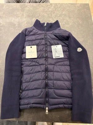 Mörkblå quiltad jacka från Moncler - Säljer en mörkblå quiltad jacka från Moncler med stickade ärmar och hög krage. Jackan har dragkedja framtill och diskret Moncler-logga på ärmen. Materialmix med dunfyllning i kroppen och stickad ull på ärmarna ger en snygg kontrast.