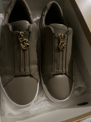 Beige sneakers från Michael Kors - Snygga beige sneakers från Michael Kors med guldfärgade dragkedjor och MK-logga framtill. Skorna har vit sula och diskret MK-mönster på sidorna. Tillverkade i skinnliknande material och har en rund tå. Perfekta för dig som gillar stilrena detaljer.