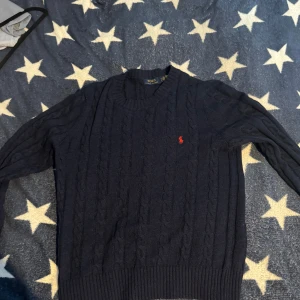Ralph lauren stickad tröja  - Klassisk mörkblå kabelstickad tröja från Polo Ralph Lauren med rund halsringning och den ikoniska röda logon broderad på bröstet. Tröjan har ribbade muddar och är långärmad, perfekt för lager på lager under kyliga dagar.