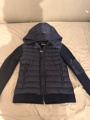 Mörkblå cardigan med huva från Monclerr - Snygg mörkblå dunväst från Monclerr med stickade ärmar och quiltad kropp. Västen har huva, dragkedja framtill, fickor med knapp och dragkedja samt en Monclerr-logga på ärmen. Perfekt för kyliga dagar och riktigt clean look. Passar mig som är 178cm