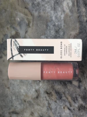 Fenty Beauty Gloss Bomb Fussy 02 - Fenty Beauty Gloss Bomb i färgen Fussy 02 är ett läppglans med en mjuk rosa ton och subtilt skimmer. Kommer i en stilren, genomskinlig tub med ljusrosa lock och rymmer 2 ml. Ger glansiga, fylliga läppar och har en smidig applikator.