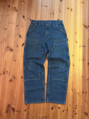 Carhartt Double Knee Pants - Säljer ett par klassiska blå byxor från Carhartt i slitstark jeans med raka ben och flera stora fickor fram och bak. Byxorna har hällor för bälte och en praktisk verktygsficka på sidan. Perfekta för dig som gillar workwear-stil.