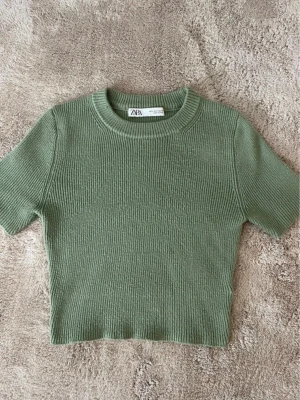 Grön ribbad topp från Zara - Säljer en grön ribbad topp från Zara i storlek S. Toppen har korta ärmar, rund halsringning och är croppad i modellen. Perfekt till jeans eller kjol för en enkel men snygg look.