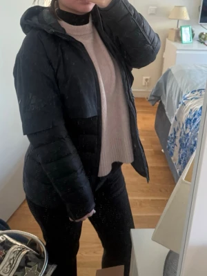 Svart pufferjacka med huva adidas - Säljer en svart pufferjacka från adidas med huva och dragkedja framtill. Jackan har quiltad design på nedre delen och slät ovandel, samt två praktiska fickor med dragkedja. Perfekt för kalla dagar och riktigt skön att bära.