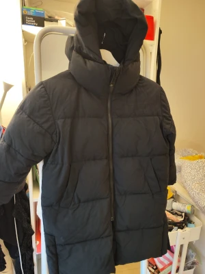 Svart pufferjacka från Molo, strl 116 - Säljer en svart pufferjacka från Molo i storlek 116. Jackan har en hög krage, huva och dragkedja framtill. Insidan är fodrad i en snygg gul färg. Perfekt för kalla dagar och riktigt skön att bära. Ny jacka kan kosta ca. 1200kr.