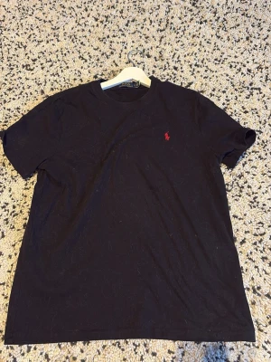 Svart t-shirt från Polo Ralph Lauren - Klassisk svart t-shirt från Polo Ralph Lauren med röd broderad logga på bröstet. T-shirten har rund halsringning och korta ärmar. Tillverkad i mjuk bomull för skön känsla och enkel stil. Tröjan är för tillfället lite hårig men det fixar jag vid köp så blir den som ny 