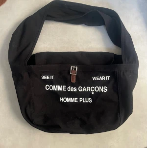 Vintage Comme des Garçons Homme Plus axelväska  - Säljer en svart axelväska från Comme des Garçons Homme Plus med vit text på framsidan och ett spänne i brunt läder. Väskan har ett stort fack, innerficka med dragkedja och bred axelrem. Perfekt för dig som vill ha en stilren och unik väska.