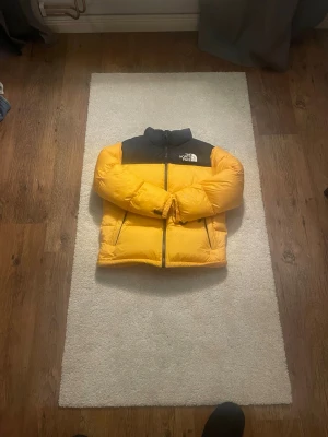 The North Face Nuptse pufferjacka - Säljer en ikonisk gul och svart The North Face 1996 Retro Nuptse pufferjacka i storlek XL Youth. Jackan har hög krage, dragkedja framtill, två sidofickor och klassisk boxig passform. Perfekt för kalla dagar och riktigt snygg streetstil.