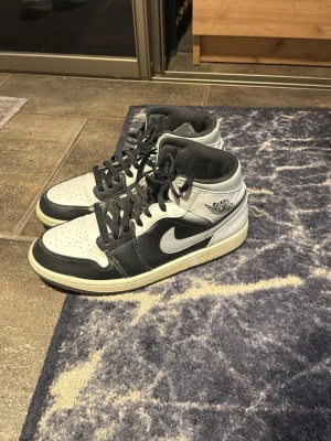 Nike Air Jordan 1 svart/vit mid - Säljer ett par klassiska Nike Air Jordan 1 high sneakers i svart och vitt skinn. Skorna har svart snörning, vit sula och den ikoniska swoosh-loggan på sidan. Perfekta för dig som gillar streetwear och vill ha en tidlös look.EJ BOX