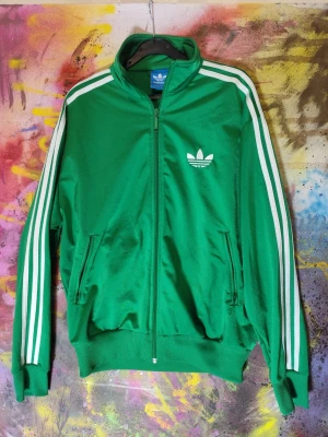 Adidas Originals grön track jacket – zip – storlek M - Klassisk Adidas Originals track jacket i grön färg med vita ränder och trefoil-logga. Storlek M polyester. Klassisk track jacket med två fickor med fungerande dragkedja.  Det finns mindre ytliga bruksspår/ruggning i tyget på ett ställe (se bild). Inget som påverkar användning eller helhetsintryck.  Hör av dig vid frågor.