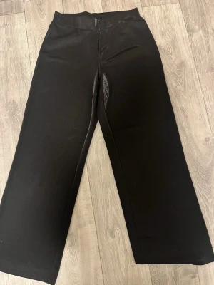 Svarta vida byxor med glans - Säljer ett par svarta byxor från Gina Tricot i storlek M. Byxorna har en hög midja och ett snyggt wide fit-ben. Materialet är glasigt. Perfekta att styla med en crop top eller oversized hoodie. Byxorna har aldrig använts 