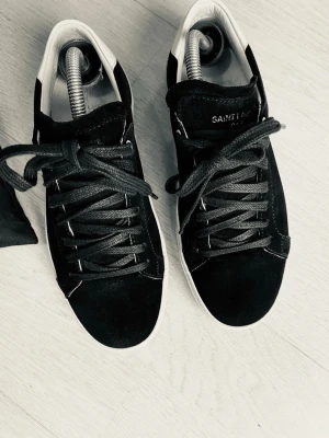 Svarta sneakers från Saint Laurent - Säljer ett par svarta sneakers från Saint Laurent Paris i mocka och skinn med vita detaljer och klassisk snörning. Snygg och stilren design med vit sula och svart ovandel. Skorna är tillverkade i Spanien och kommer med original dustbag.