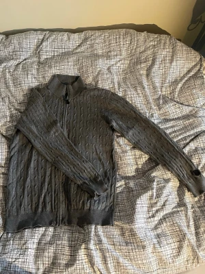 Grå kabelstickad kofta Cutter & Buck - Säljer en grå kabelstickad kofta från Cutter & Buck i storlek M eftersom den blivit för liten. Snygg detalj med logomärke på ärmen. Perfekt för lager på lager under kyliga dagar eller som en tjockare tröja på en sommardag. Hör av dig vid fundering.