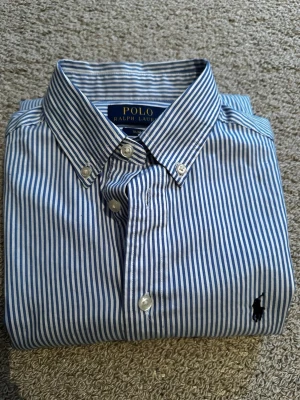 Oxford skjorta från Polo Ralph Lauren - Säljer en blåvit randig skjorta från Polo Ralph Lauren i slim fit-modell. Skjortan har klassisk button-down krage, långa ärmar och broderad logga på bröstet. Tillverkad i mjuk bomull och har knappar framtill. Sitter som xs skulle jag säga 