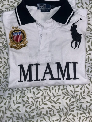 Vit Polo Ralph Lauren skjorta MIAMI - Säljer en vit Polo Ralph Lauren skjorta med svart krage och stora broderade detaljer. Framsidan har MIAMI-text, klubbmärke och den klassiska polospelaren. Baksidan har en stor svart siffra 5. Skjortan är i bomull och har korta ärmar.
