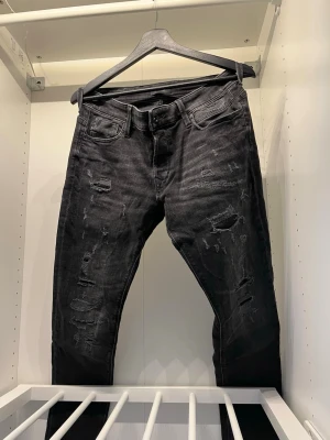 Svarta slitna skinny jeans - Säljer ett par svarta skinny jeans med slitningar och trasiga detaljer på både fram- och baksida. Jeansen har klassisk femficksmodell och knappgylf. Perfekta för dig som gillar en edgy och trendig stil.