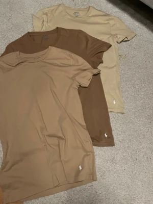 Beige och bruna t-shirts Polo Ralph Lauren - Tre stilrena t-shirts från Polo Ralph Lauren i olika beige och bruna nyanser. Klassisk passform med korta ärmar och rund hals. Diskret broderad logga vid nederkanten. Perfekta basplagg till garderoben och tillverkade i mjuk bomull.