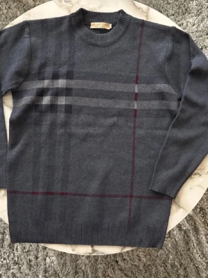 Grå rutig tröja från Burberry - Säljer en klassisk grå tröja från Burberry med rutigt mönster i mörkgrått och vinrött. Tröjan har rund halsringning och långa ärmar. Perfekt för dig som gillar stilrena och tidlösa plagg.