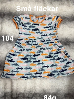  klänning med biltryck - Söt klänning med färgglatt biltryck i blått, orange och rosa. Klänningen har orange kantband vid ärmar och hals, samt korta ärmar. Perfekt för dig som gillar lekfulla mönster och bekväma plagg.🌸 Små fläckar. Storlek 104