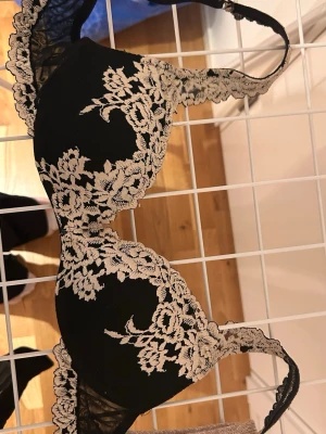 Svart spets-bh med blommig spets - Snygg svart bh med beige blommig spetsdetalj över kuporna och längs banden. Bh:n har tunna axelband och transparent mesh på sidorna. Perfekt för dig som gillar en elegant och romantisk look. Använd endast en gång.