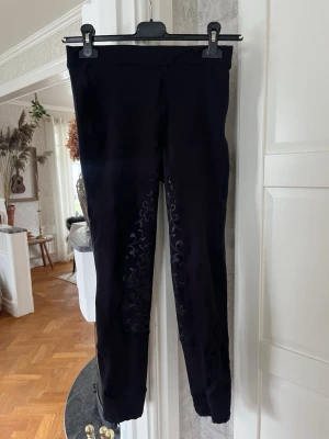 Svarta ridleggings med mönster, stl 150 - Säljer ett par svarta rid byxor i storlek 150. De har snygga blommiga detaljer på insidan av benen och bakdelen. Tillverkade i 100% polyester och har en tight passform som sitter skönt vid ridning. Har ett litet hål i skrevet men går att sy ihop för den som orkar!