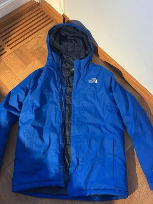 Blå vindjacka från The North Face  - Säljer en blå vindjacka från The North Face. Jackan har huva, dragkedja framtill och två sidofickor. Perfekt för blåsiga dagar och enkel att matcha med olika outfits. Klassisk logga på bröstet och ryggen.