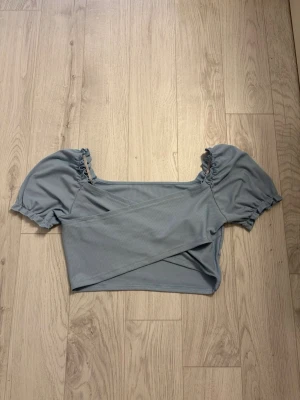 Ljusblå omlott topp med puffärm - Säljer en ljusblå croppad topp med omlottdesign och puffiga korta ärmar. Toppen har en ribbad struktur och resår vid ärmsluten för en söt look. Perfekt till jeans eller kjol för en trendig outfit.