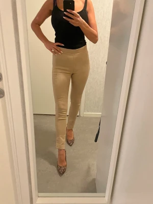 Beiga tighta byxor med slits - Säljer ett par beiga, högmidjade byxor i stretchigt material. Byxorna har en tight passform och slits nertill vid ankeln för en snygg detalj. Perfekta att styla med klackar eller sneakers. Supersköna och smickrande modell. MNG Suit