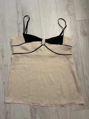 Beige och svart linnetopp från Mango - Säljer en snygg beige och svart linnetopp från Mango med tunna axelband och kontrasterande svarta detaljer. Toppen har en stilren design med markerad byst och mjukt, glansigt material som ger en lyxig känsla.