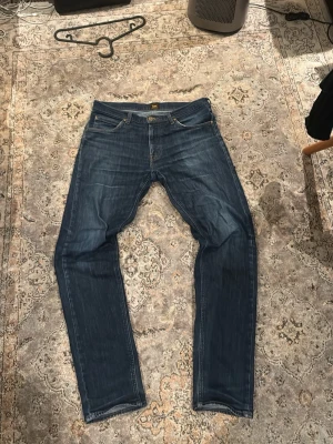 Mörkblå Lee jeans straight fit - Säljer ett par mörkblå jeans från Lee med klassisk femficksdesign och raka ben och väldigt fin wash. Jeansen har Lee-lapp bak i midjan och snygga slitningar framtill för en avslappnad look. Tillverkade i slitstarkt denimtyg som passar perfekt till vardagsstilen. Storlek 34 men sitter mer som 32-33