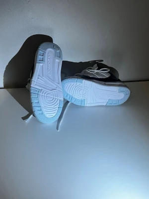 Svarta och vita Jordan sneakers - Säljer ett par svarta och vita Jordan sneakers med högt skaft och vita snören. Skorna har en blank svart tå och vit sula med tydlig Jordan-logga. Ovandelen är i textil och syntet, och de har en klassisk sportig look.