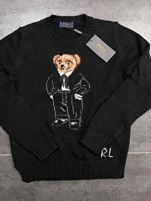 Svart stickad tröja Polo Ralph Lauren - Svart stickad tröja från Polo Ralph Lauren med ikonisk björn i kostym broderad på bröstet. Rund halsringning och ribbade muddar vid ärmslut och nederkant. RL-broderi nedtill. Perfekt för dig som vill ha en klassisk men lekfull look.