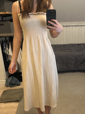 Ljusrosa långklänning från H&M - Säljer en ljusrosa långklänning från H&M med smala axelband och smock upptill. Klänningen har en luftig passform och volangkant vid bysten. Perfekt för varma dagar och enkel att styla med accessoarer. Tycker egentligen den är supersöt, men den är tyvärr för stor för mig. Har satt en hårklämma bak på bilderna