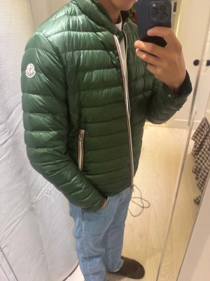 Grön Moncler rigel jacket - Snygg grön pufferjacka från Moncler i storlek  S Jackan har quiltad design, lång ärm och klassisk Moncler-logga på ärmen. Dragkedja framtill med coola rödvita detaljer och två fickor med zip. Perfekt för dig som vill ha en clean och stilren look.